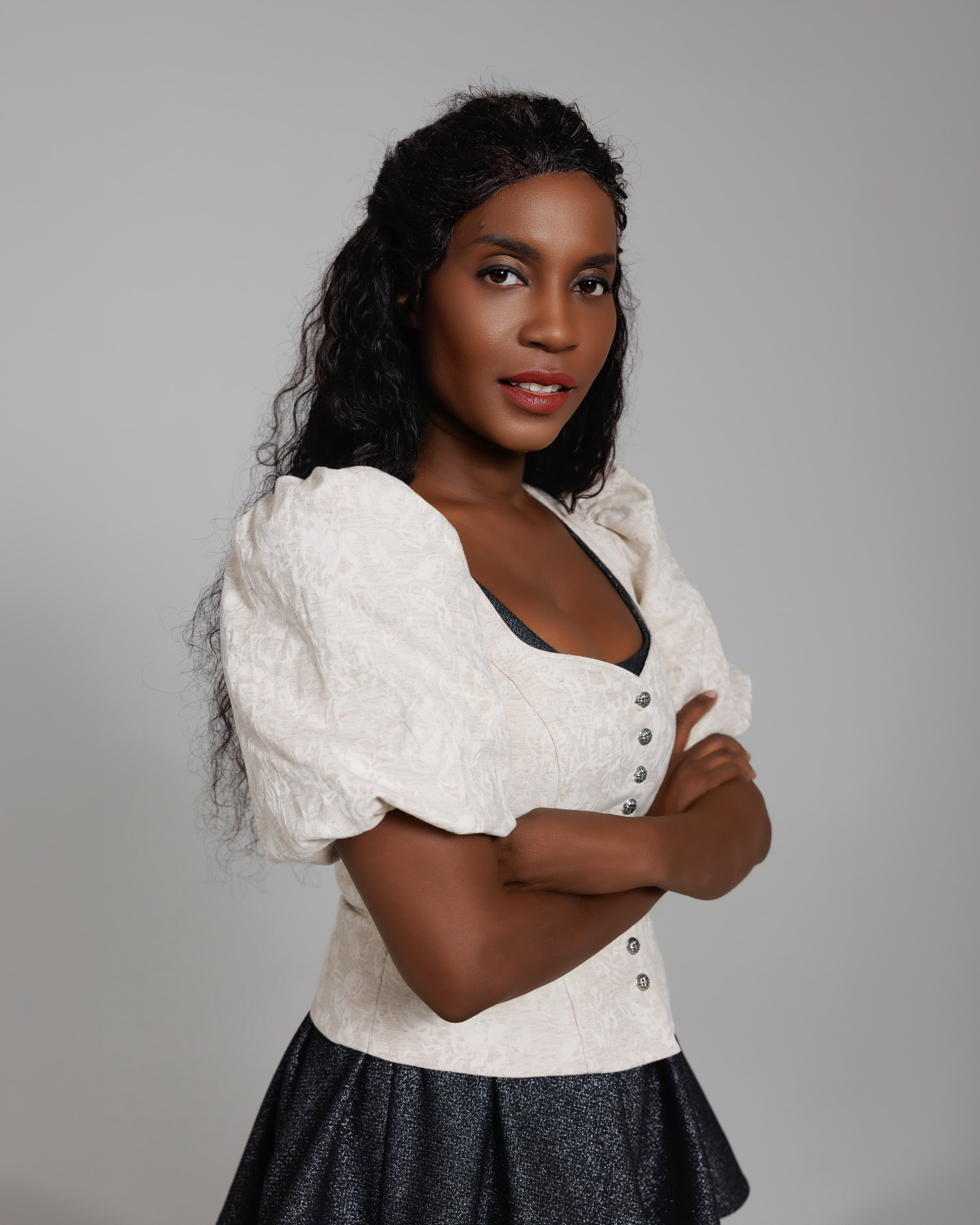 Schwarze Frau mit langen, dunklen Locken, weißer Puffärmel-Bluse und verschränkten Armen blickt in die Kamera.