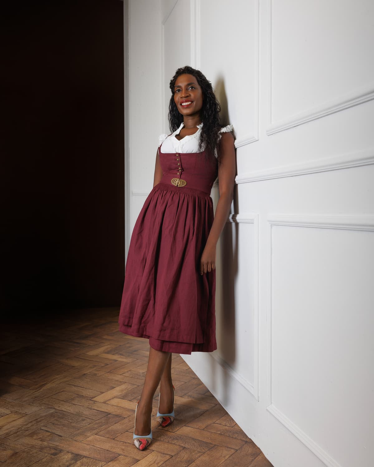 Sadia De Kiden im bordeauxroten Dirndl an eine weiße Wand gelehnt